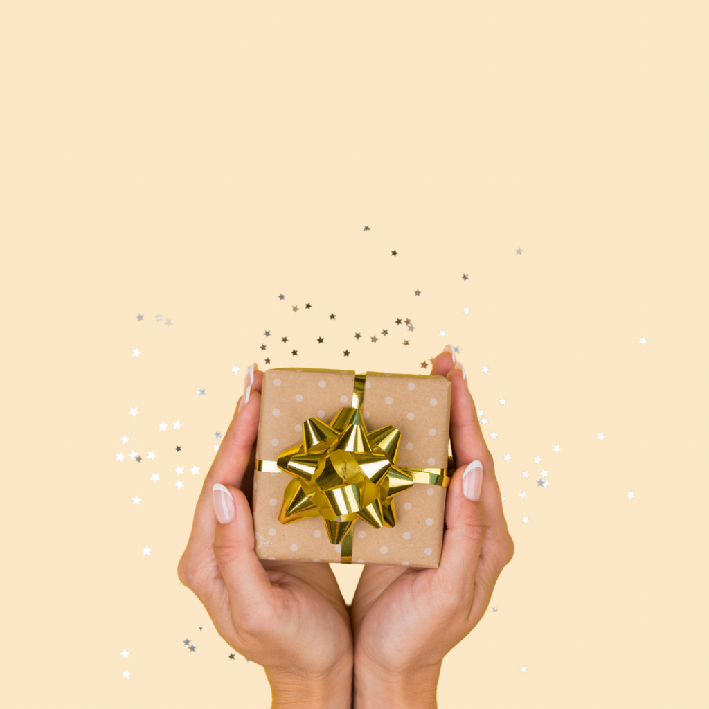 Woman’s hands holding a small polka-dotted gift box with a shiny gold bow, set against a pastel peach background with scattered confetti.