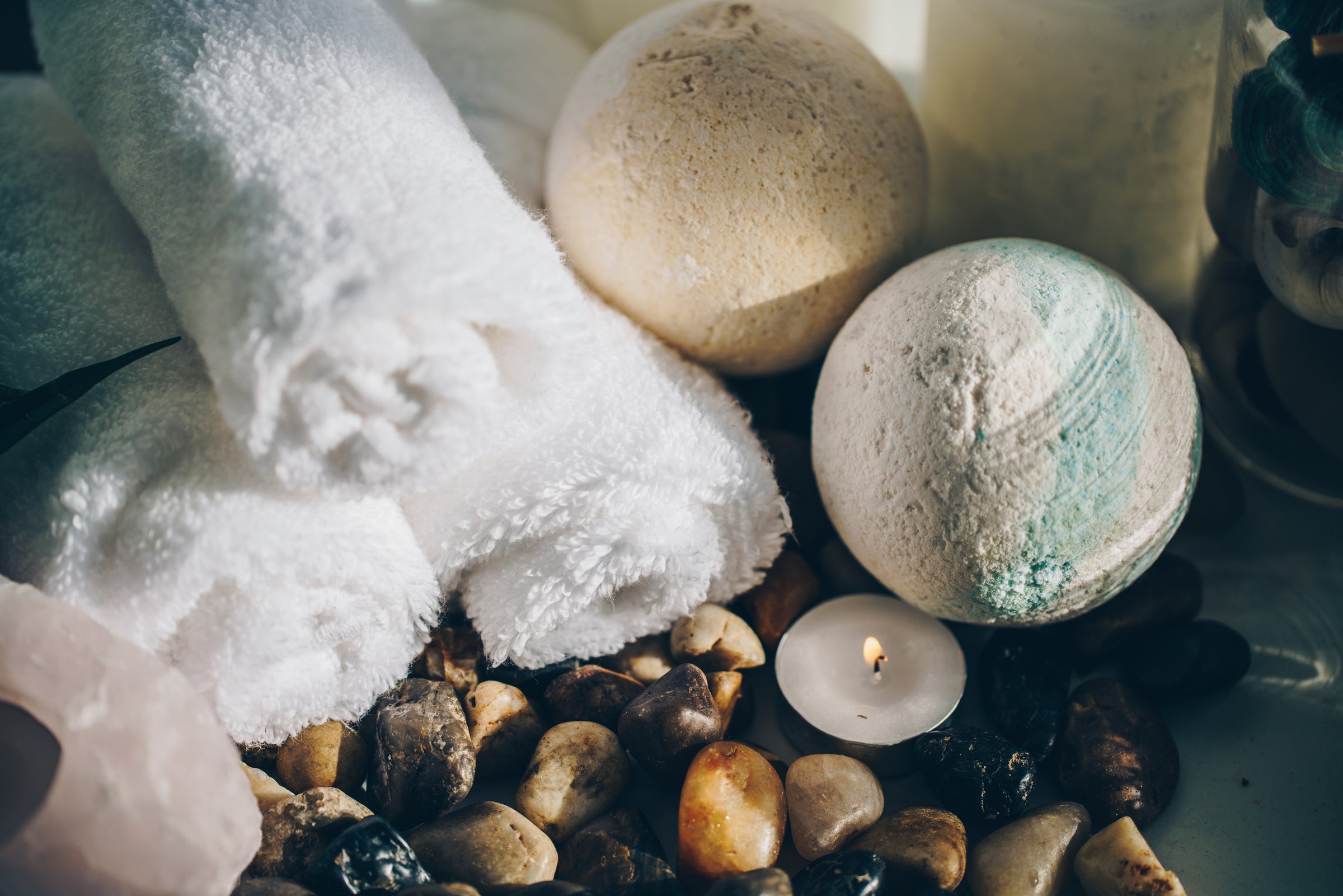 Spa-inspired flatlay with white rolled towels, bath bombs, a lit tealight candle, and smooth massage stones