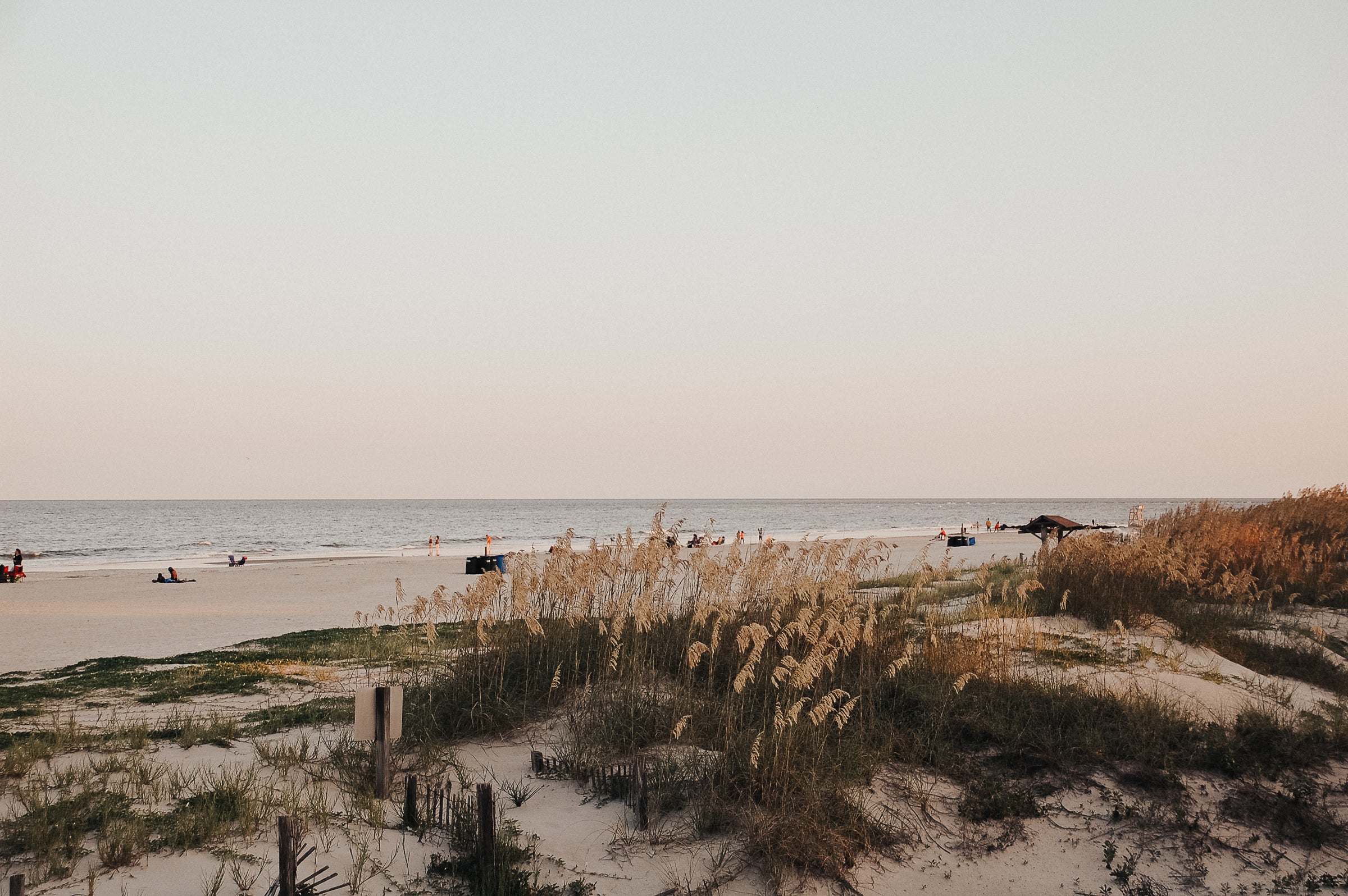 Coastal view of a serene beach during golden hour, with soft sand dunes, sea oats swaying in the breeze, and people relaxing near the shoreline under a pastel sky.