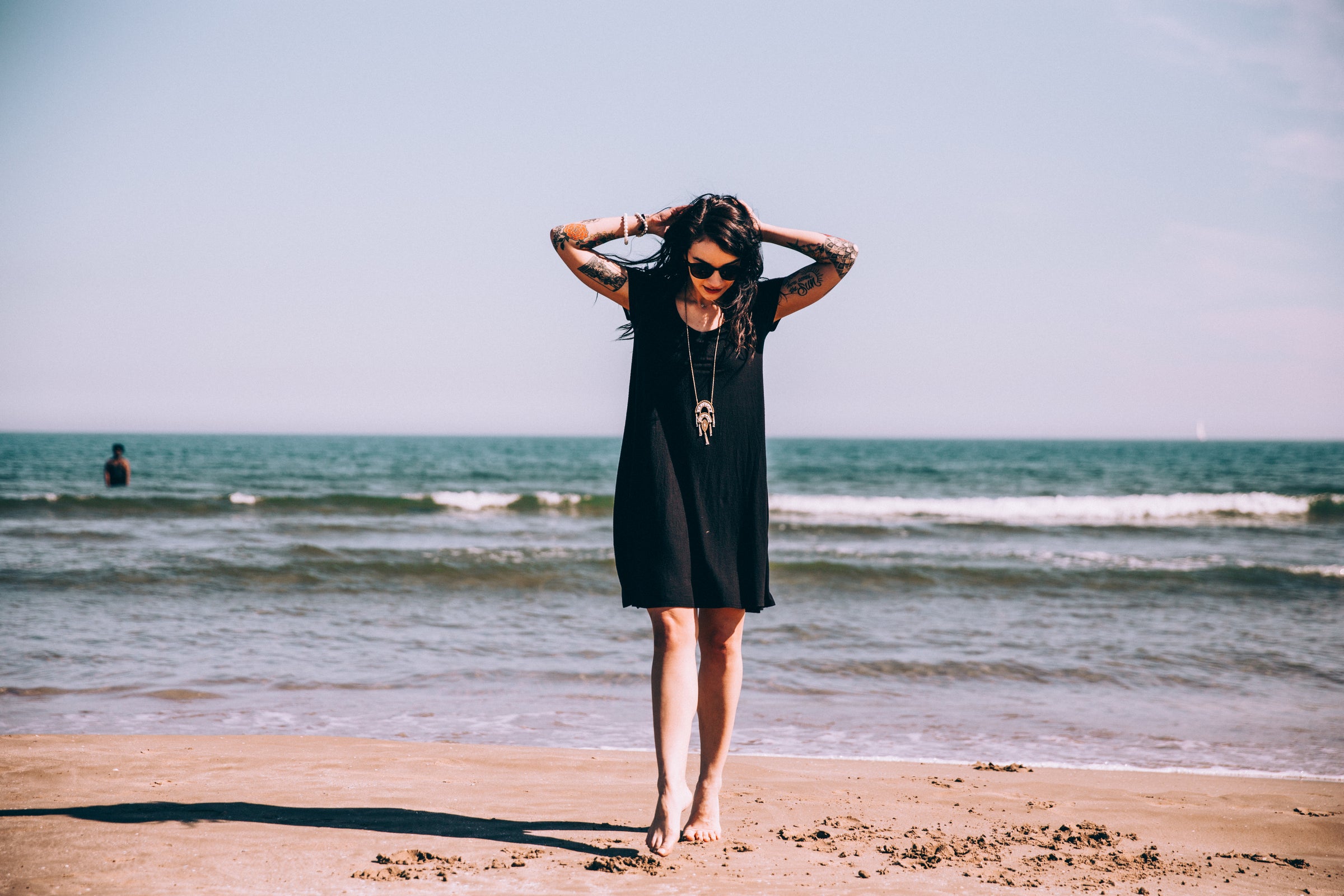 Stylish woman with tattoos walking barefoot on the beach in a black dress, enjoying the ocean breeze under a clear sky.