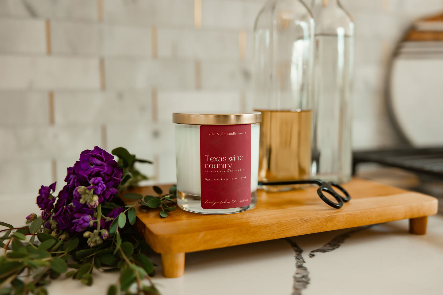 Candle with a red label on a wooden stand next to purple flowers