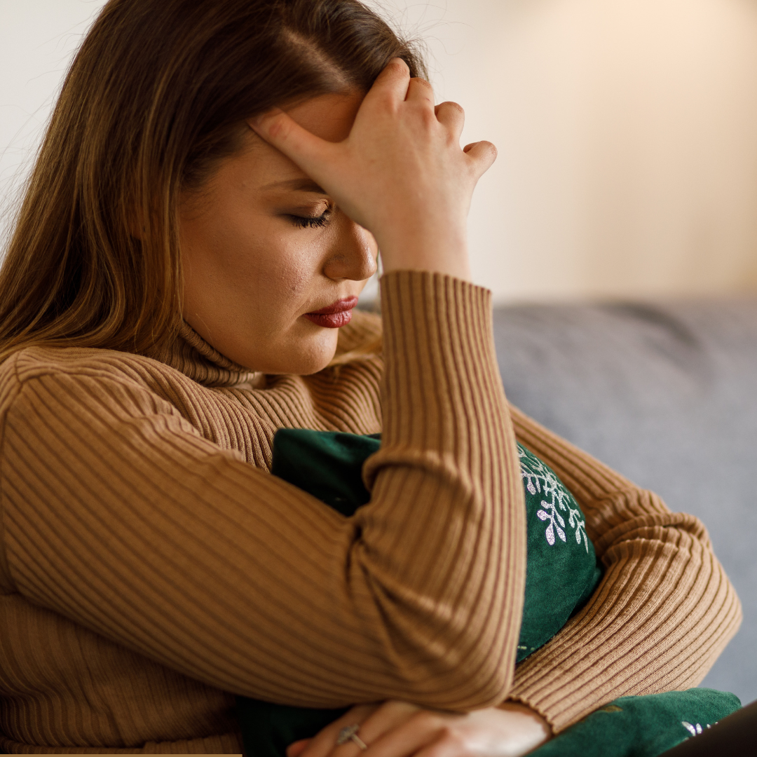 A woman sitting on a couch wearing a beige ribbed sweater, resting her head on her hand, and holding a green pillow with a snowflake design. She appears deep in thought or stressed.