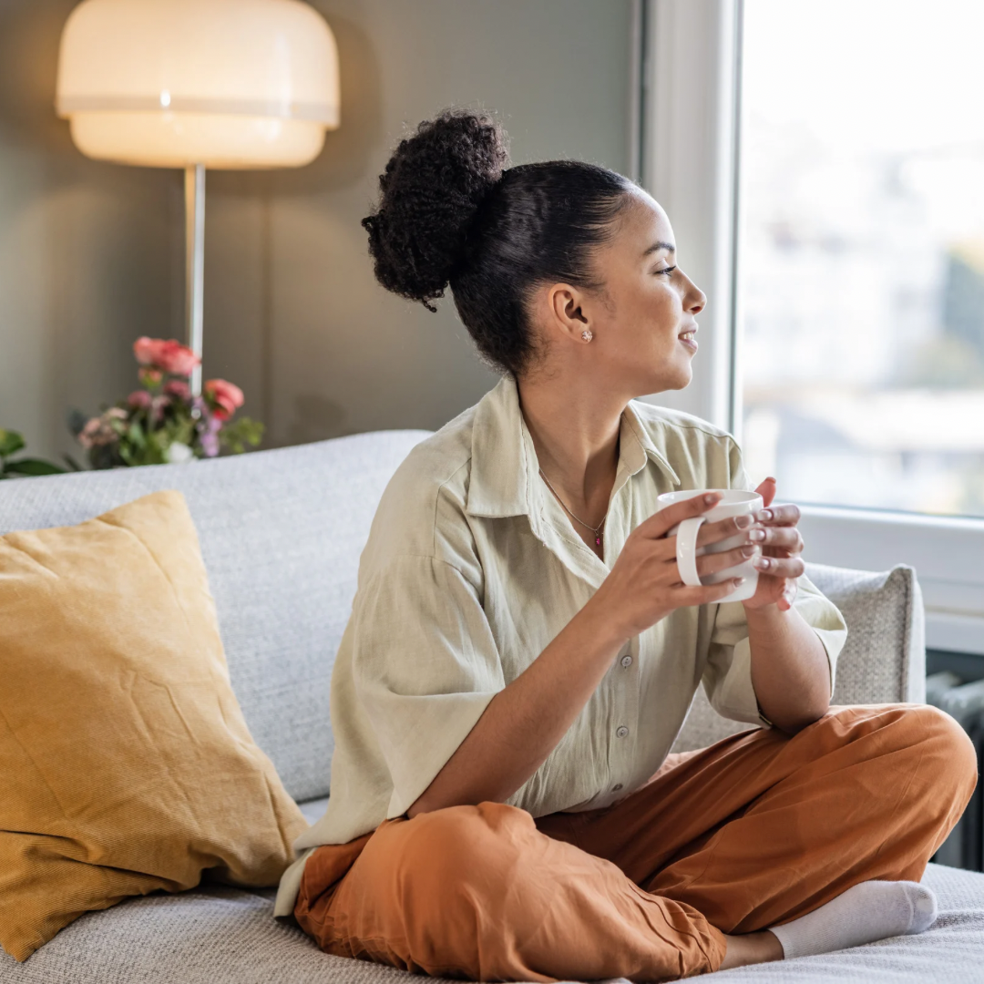 Person sitting on a couch with legs crossed, holding a white mug, gazing out of a large window. The cozy room features soft lighting, a mustard-colored pillow, and a potted plant in the background.