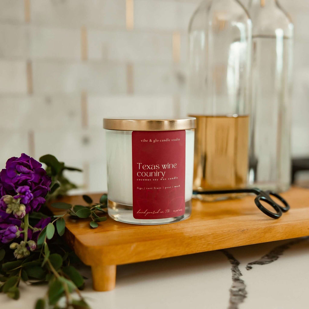 Texas Wine Country candle in sleek glass jar on wooden tray with scissors and purple flowers.