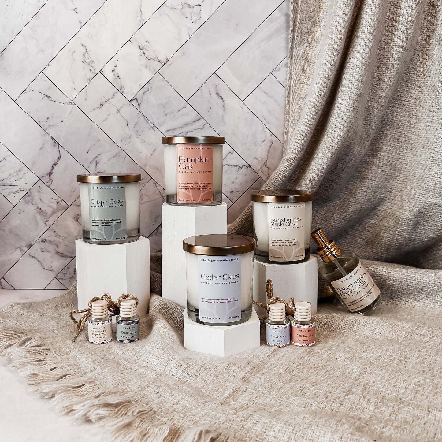 Vibe & Glo Fall 2025 Collection with four coconut soy candles, matching car diffusers, and a room + linen spray in scents Crisp + Cozy, Pumpkin + Oak, Cedar Skies, and Baked Apples + Maple Crisp, styled on pedestals with a cozy blanket backdrop.