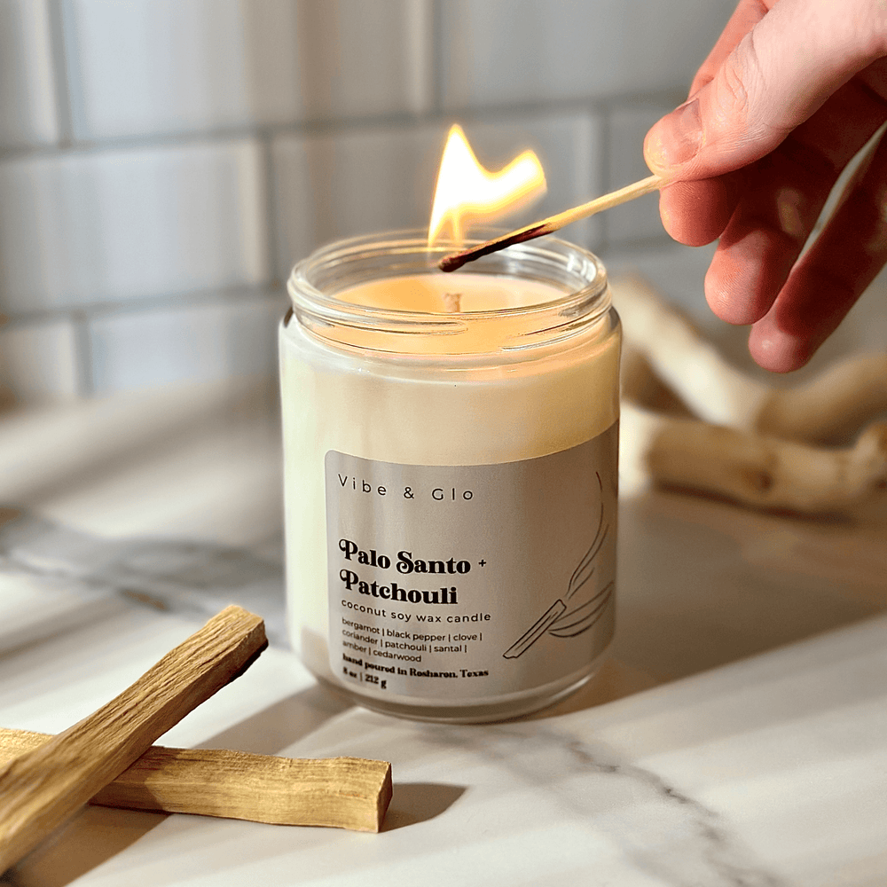 
                  
                    A lit Palo Santo + Patchouli candle from Vibe & Glo, with a matchstick lighting the wick. The candle sits on a marble surface, accompanied by pieces of palo santo wood, creating a warm and serene ambiance.
                  
                