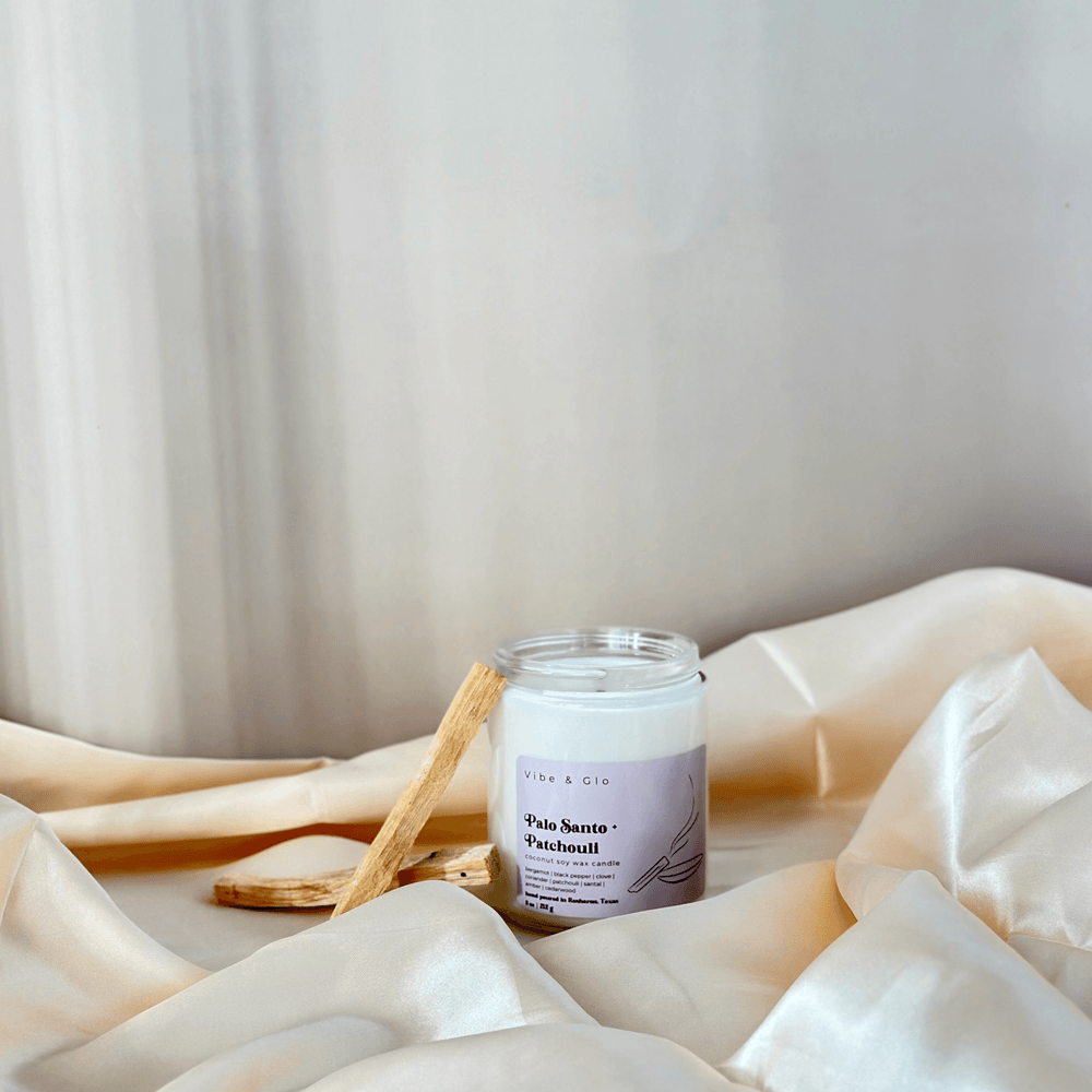 
                  
                    The Palo Santo + Patchouli candle by Vibe & Glo, unlit, resting on soft, elegant golden fabric with pieces of palo santo wood nearby. The minimalist and serene setting highlights the candle’s luxurious and eco-friendly design.
                  
                