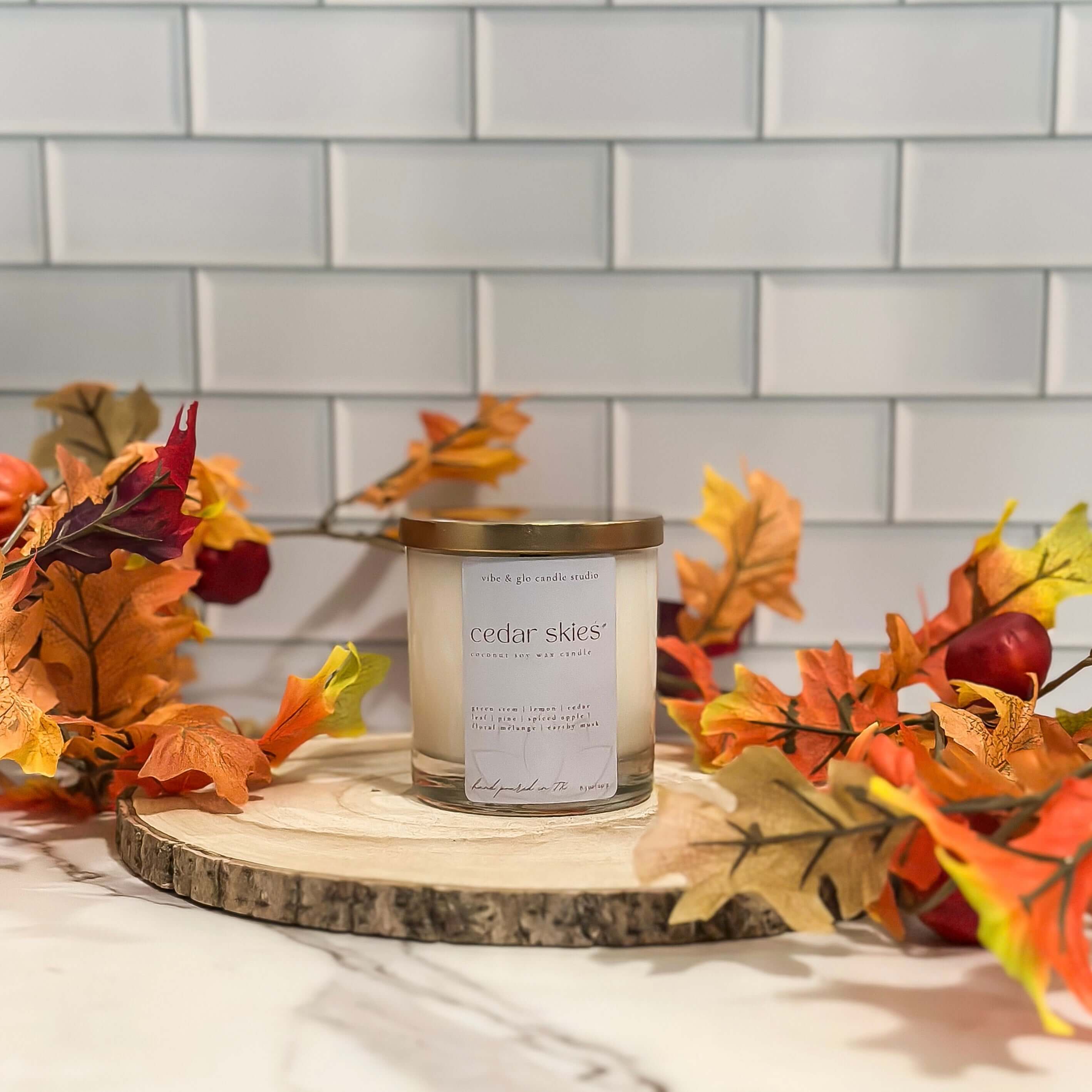 Cedar Skies coconut soy fall candle in clear glass jar with gold lid by Vibe & Glo Candle Studio, styled with autumn leaves on wood slice.