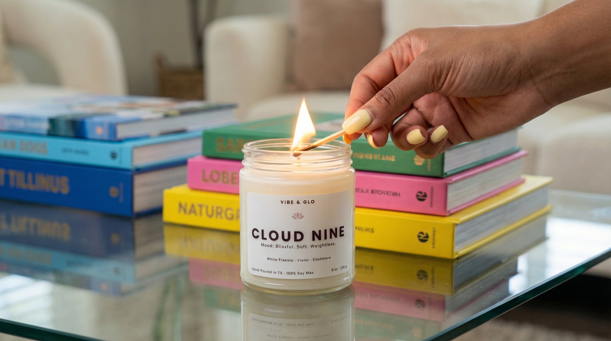 Hand lighting a Vibe & Glo Cloud Nine soy candle in a clear glass jar on a glass table, with colorful stacked books in the background creating a soft, cozy lifestyle scene.