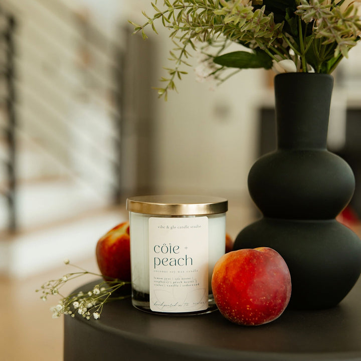 Côte + Peach candle on a table, surrounded by fresh peaches and a decorative vase with greenery.