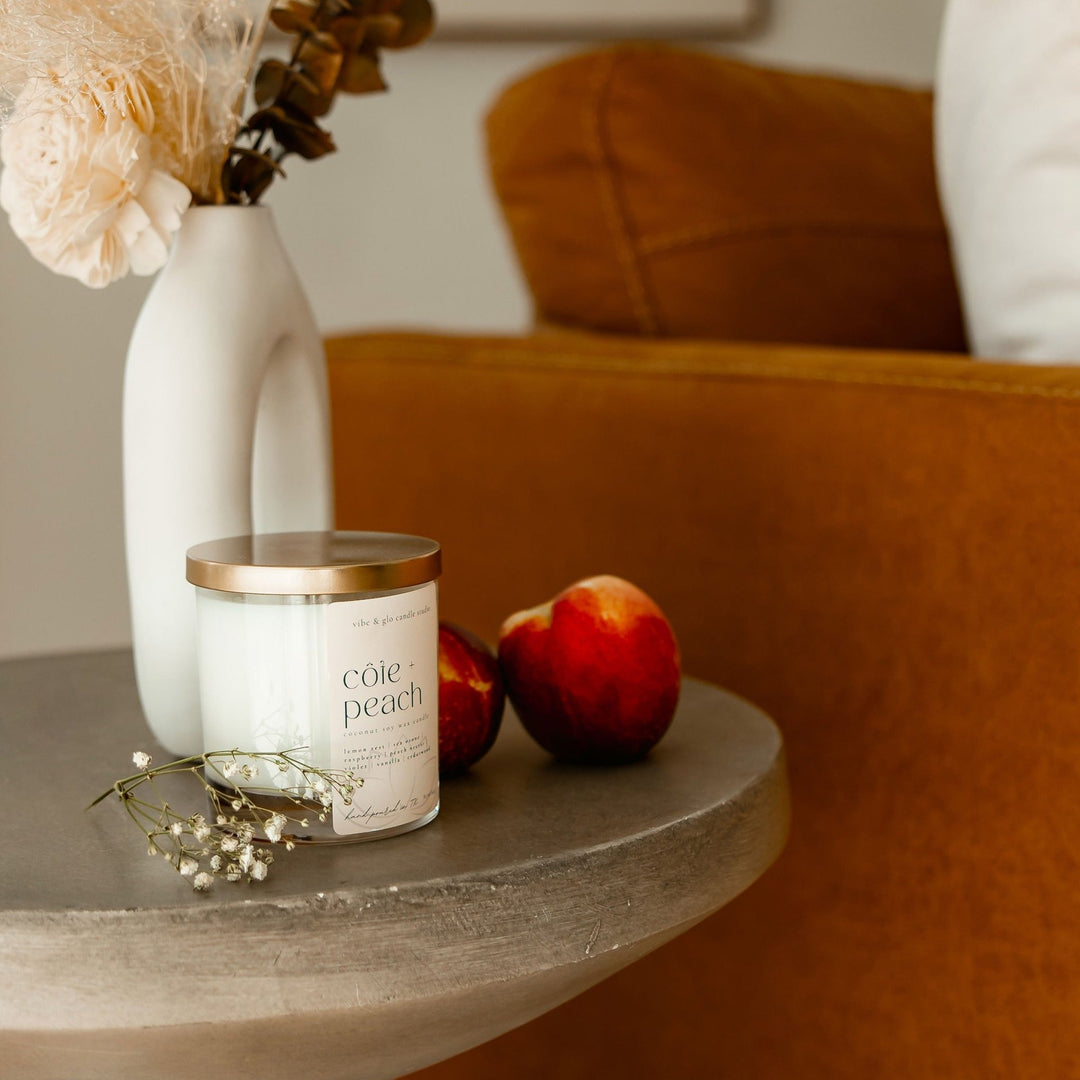 Côte + Peach candle on a table with peaches and a vase of dried flowers, capturing a coastal, sunny vibe.