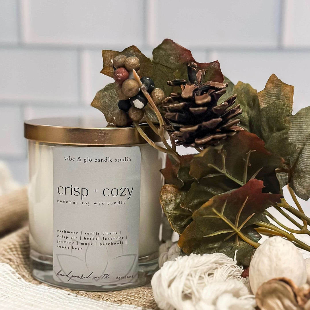 Crisp + Cozy coconut soy fall candle in white glass jar with gold lid by Vibe & Glo Candle Studio, styled with pinecone and autumn greenery.