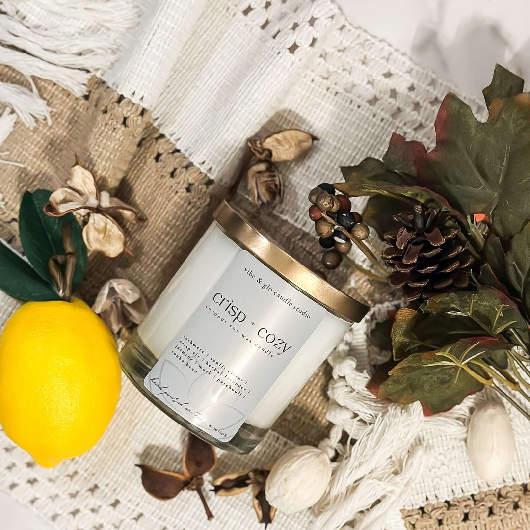Crisp + Cozy coconut soy fall candle in clear glass jar with gold lid by Vibe & Glo Candle Studio, styled with lemon, pinecone, and fall greenery.