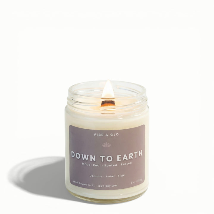 Down to Earth oakmoss, amber, and sage soy candle by Vibe & Glo with lit wooden wick in clear glass jar.