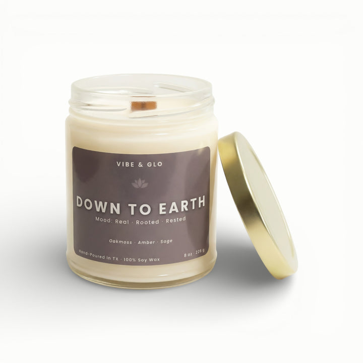 Vibe & Glo Down to Earth soy candle in clear glass jar with wooden wick and gold lid, featuring oakmoss, amber, and sage notes