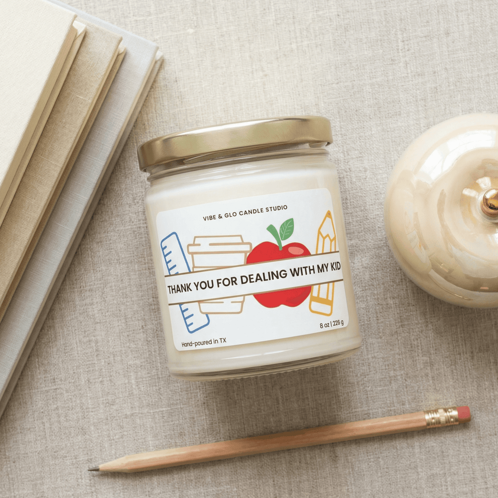 Funny teacher gift soy candle that says “Thank You for Dealing With My Kid” in 8 oz glass jar with gold lid, styled flatlay with pencil and neutral books.