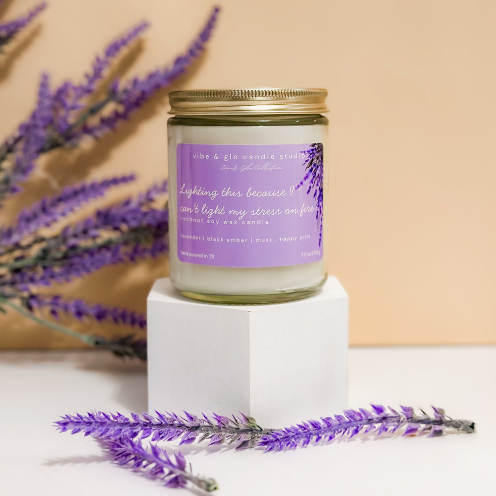 Lighting this because I can’t light my stress on fire coconut soy wax candle with lavender, black amber, and musk. Displayed on a white pedestal with lavender and peach background.