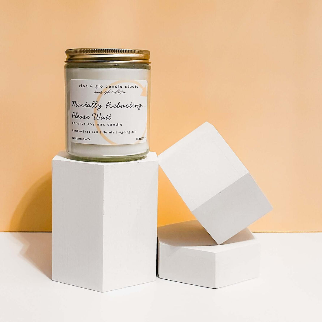 Mentally Rebooting Please Wait coconut soy wax candle with bamboo, sea salt, and floral notes. Displayed on geometric white blocks against a peach background for a modern minimal look.