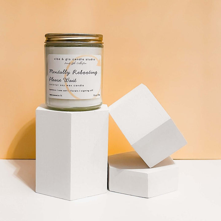 Mentally Rebooting Please Wait coconut soy wax candle with bamboo, sea salt, and floral notes. Displayed on geometric white blocks against a peach background for a modern minimal look.