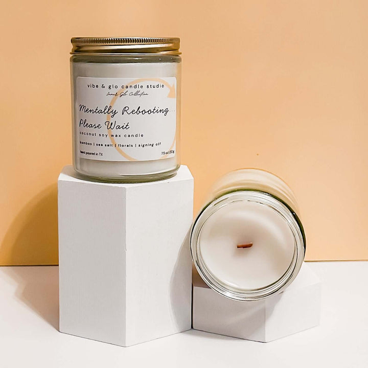  Mentally Rebooting Please Wait coconut soy wax candle with bamboo, sea salt, and floral notes. Displayed on white pedestals against a peach backdrop for a calm and modern aesthetic.