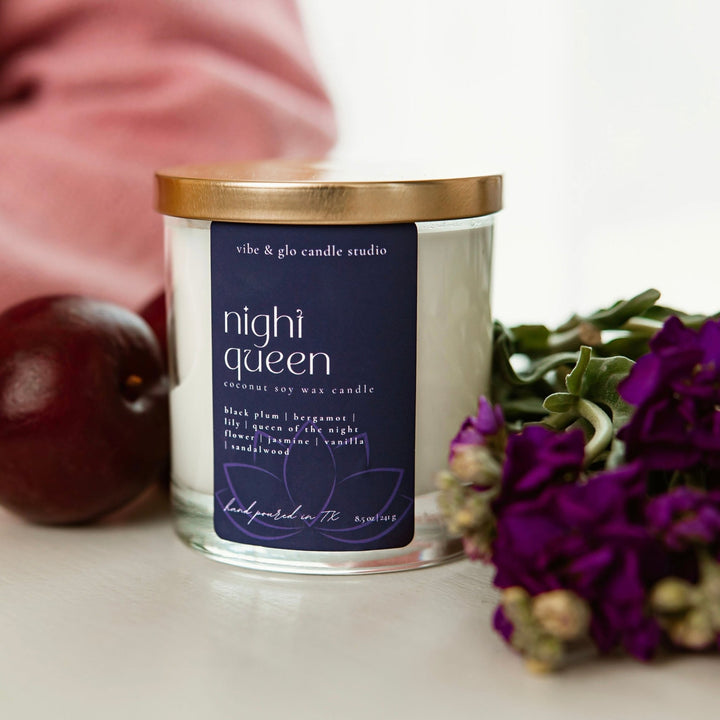 Vibe & Glo Night Queen candle in a clear glass jar with a gold lid, surrounded by purple flowers and a plum on a white table.