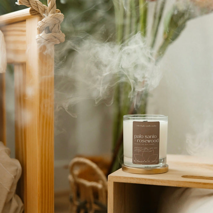 Palo Santo + Rosewood candle with curling smoke in a serene bedroom scene with natural wood decor.