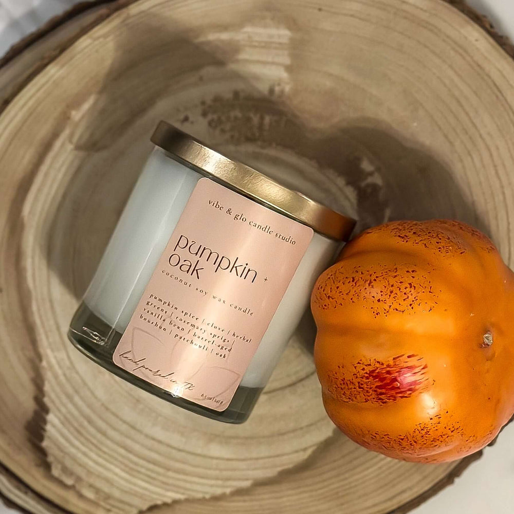 Flatlay of Pumpkin + Oak coconut soy fall candle in clear  glass jar with gold lid by Vibe & Glo Candle Studio, styled with small orange pumpkin.