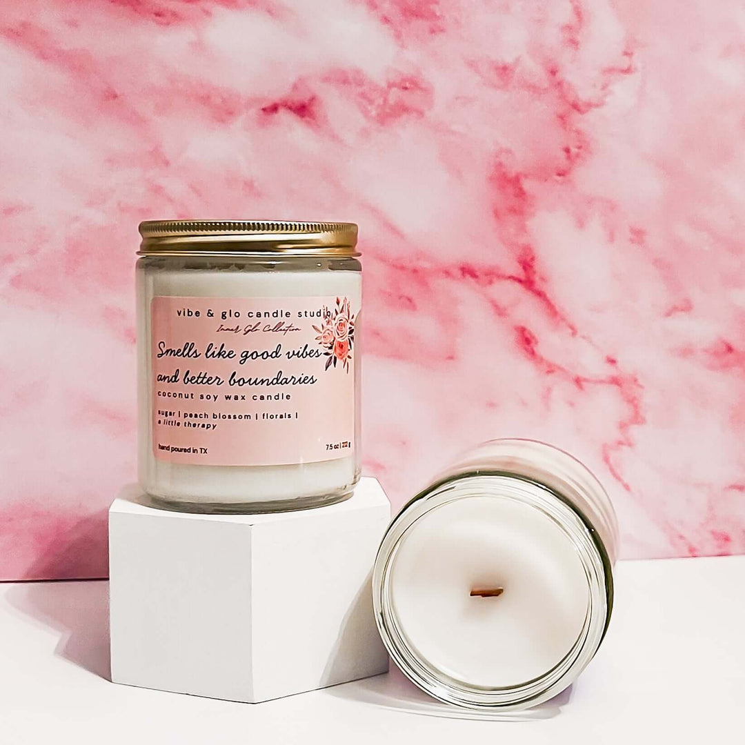  Smells like good vibes and better boundaries coconut soy wax candle with sugar, peach blossom, and florals. Displayed on a pedestal with a pink marble backdrop.