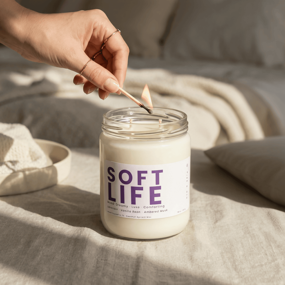 Soft Life 2-wick candle being lit on soft linen bedding, cozy relaxing home fragrance with lavender and vanilla notes