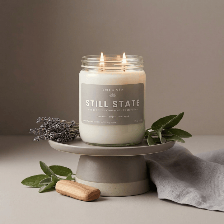 Lit Still State double wick soy candle with lavender and sage accents displayed on a ceramic pedestal, creating a soft, calming home fragrance scene.