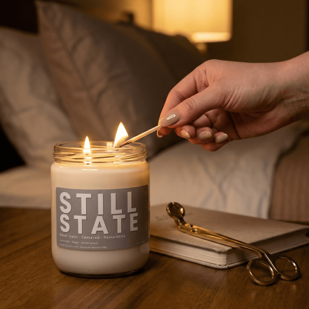 Still State 2-wick candle being lit with a match in a cozy bedroom setting, creating a warm, calming atmosphere