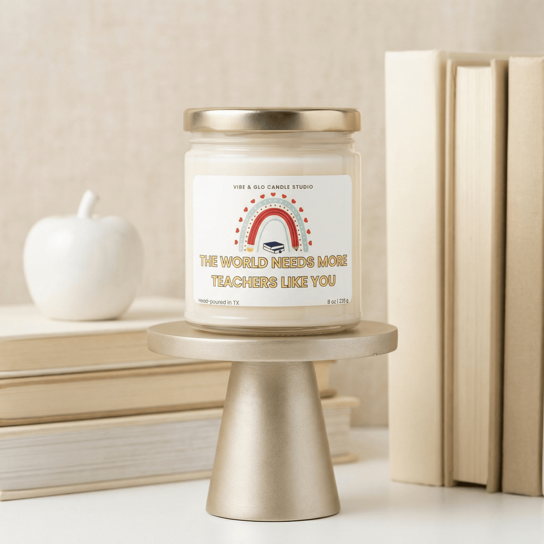 Teacher appreciation gift candle reading “The World Needs More Teachers Like You” displayed on a gold pedestal with stacked books and a white apple, 8 oz hand-poured soy candle by Vibe & Glo Candle Studio.