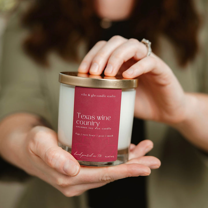 Close-up of a woman holding the Texas Wine Country candle by Vibe & Glo Candle Studio, featuring a burgundy label and gold lid.