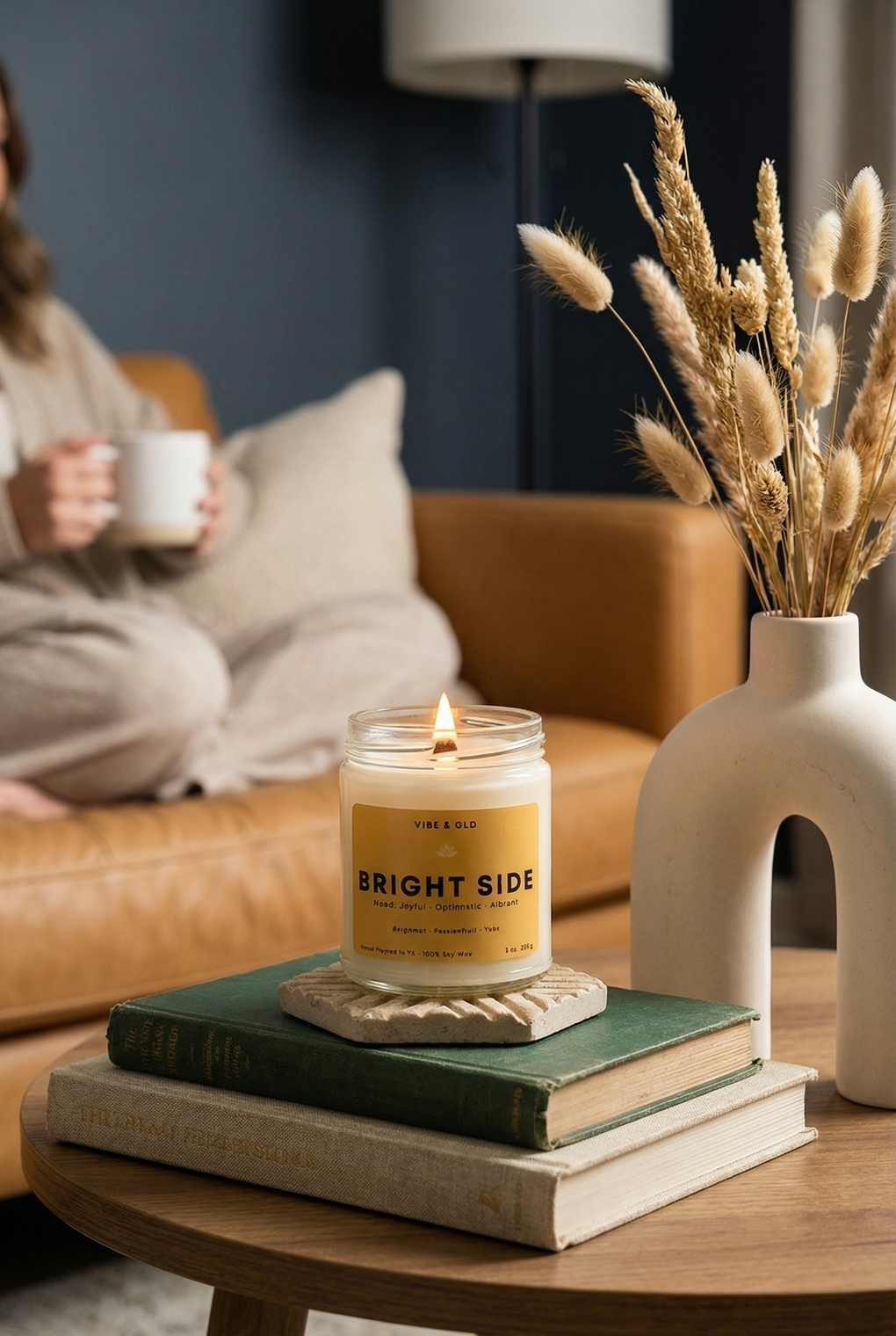 Candle labeled 'Bright Side' on books with a vase of dried plants and a person holding a mug in the background.