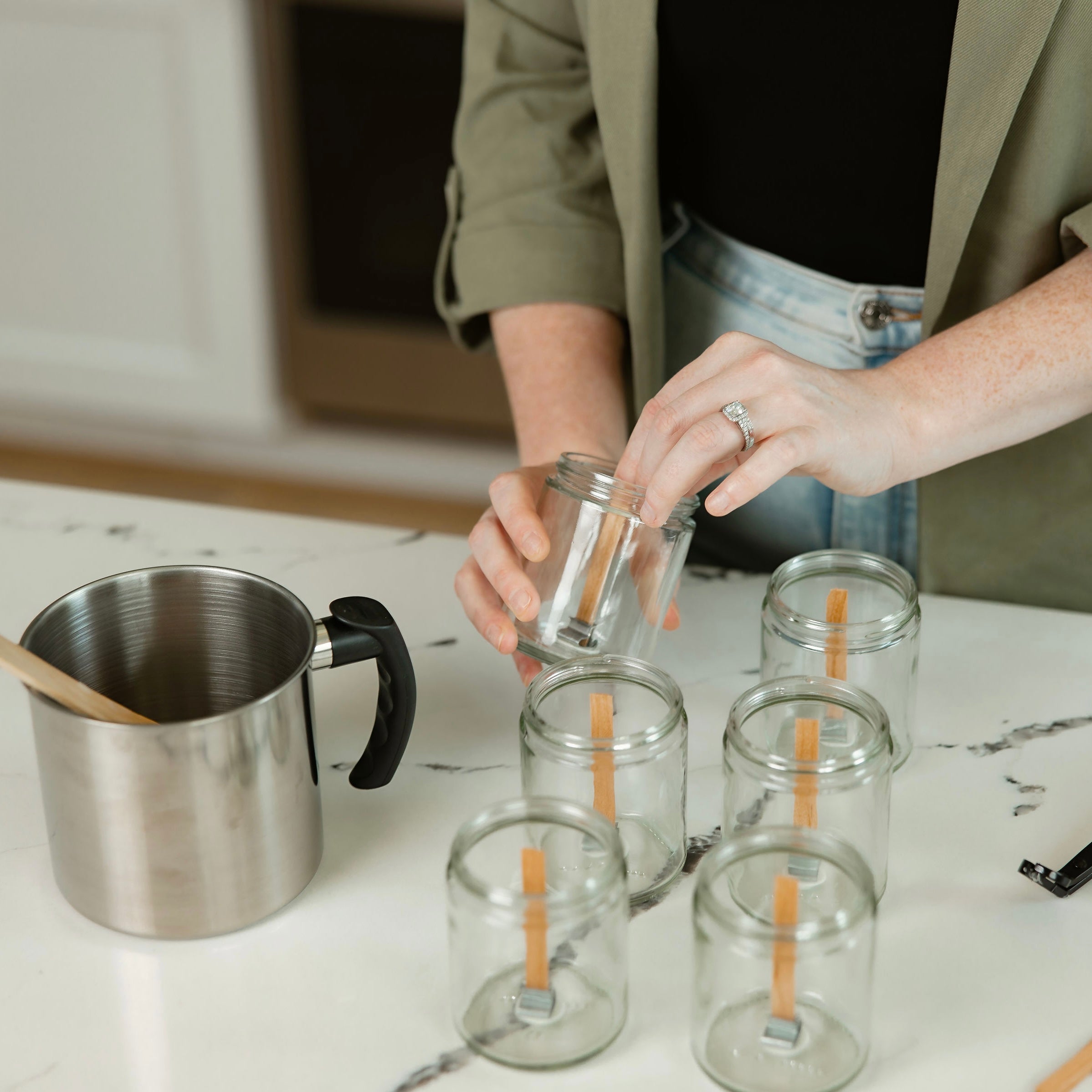 Woman prepping empty candle jars with wooden wicks in a clean kitchen workspace during the candle-making process