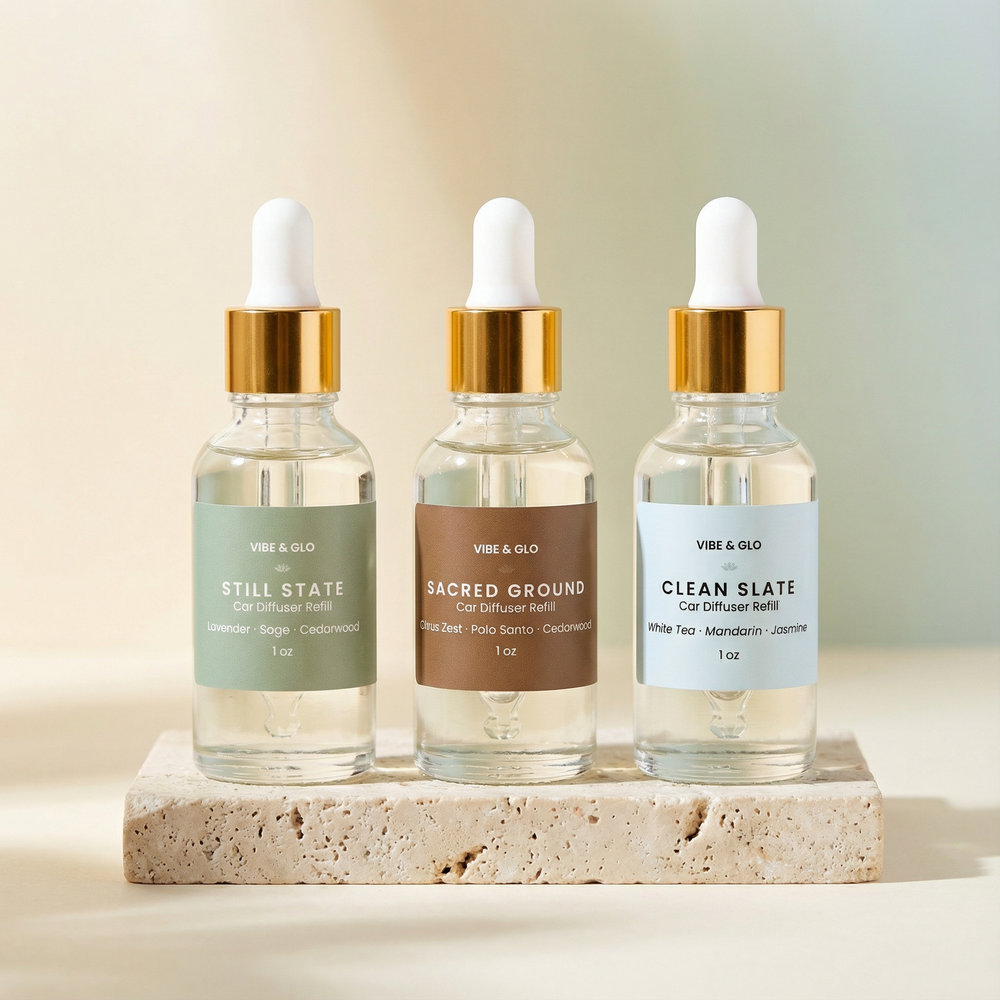 Three Vibe & Glo car diffuser refills displayed on a neutral stone pedestal, featuring clear glass bottles with visible fragrance oil and gold droppers in the scents Still State, Sacred Ground, and Clean Slate.