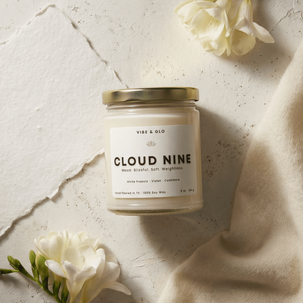 Vibe & Glo Cloud Nine soy candle with white freesia, violet, and cashmere styled with soft white florals and neutral textures in an airy flatlay.