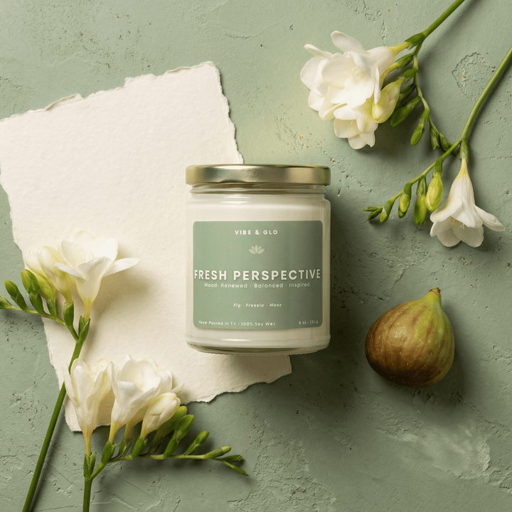 Fresh Perspective 8 oz soy candle by Vibe & Glo featuring fig, freesia, and moss surrounded by white freesia flowers and whole fig fruit in a botanical green flatlay.