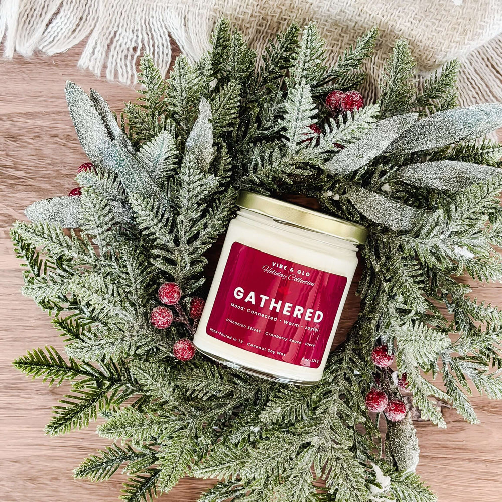 Vibe & Glo Gathered candle surrounded by a frosted wreath with red berries, showcasing coconut soy wax and festive holiday packaging.