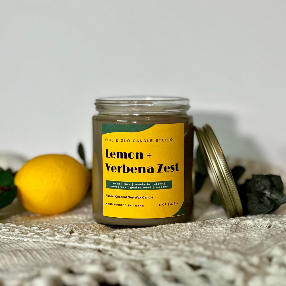 
                  
                    Vibe & Glo Candle Studio's Lemon + Verbena Zest candle with the lid off, styled with a fresh lemon on a white fabric. Features citrus and herbal fragrance notes, hand-poured in Texas.
                  
                