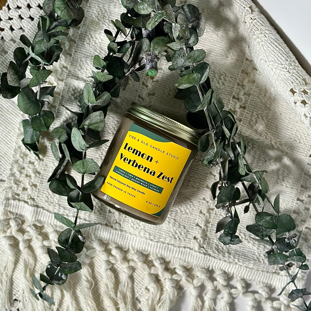 
                  
                    Vibe & Glo Candle Studio's Lemon + Verbena Zest candle surrounded by fresh eucalyptus, styled on a textured white fabric. Features a yellow and green label with bright citrus and herbal notes.
                  
                