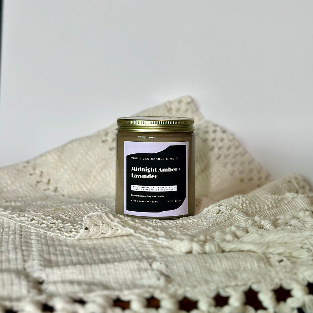 
                  
                    Vibe & Glo Candle Studio's Midnight Amber + Lavender candle displayed on a cozy white fabric. Features a bold black and white label with relaxing and floral fragrance notes.
                  
                