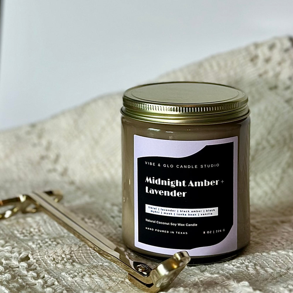 
                  
                    Vibe & Glo Candle Studio's Midnight Amber + Lavender candle styled with a gold wick trimmer on a textured white fabric. The elegant black and white label highlights floral and musky scent notes.
                  
                