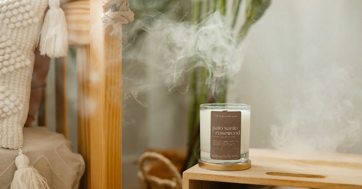 A Palo Santo + Rosewood candle by Vibe & Glo Candle Studio placed on a bohemian style wood table next to a cozy boho themed bed. There is smoke rising from the candle as if it has just been blown out.