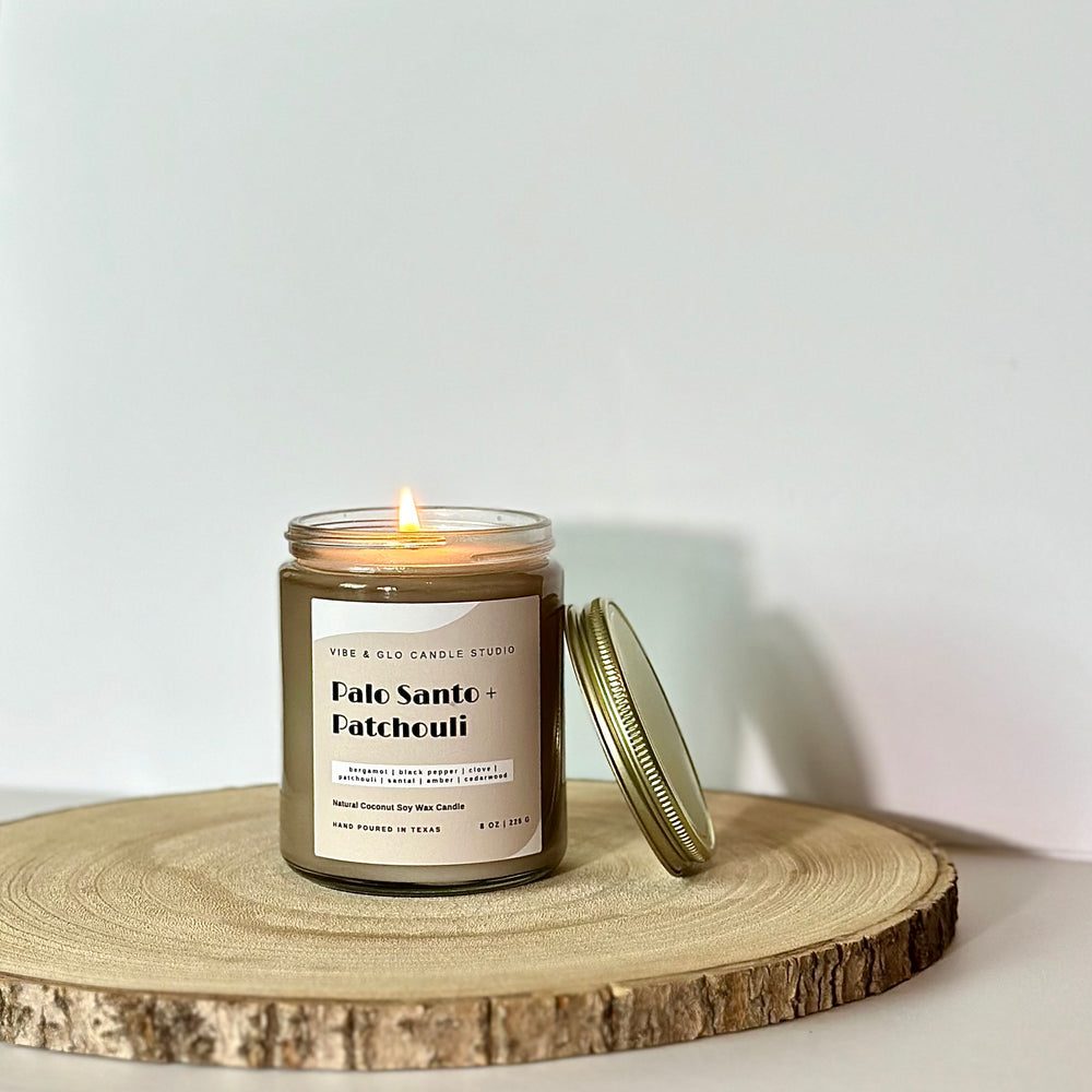 
                  
                    Vibe & Glo Candle Studio's Palo Santo + Patchouli candle lit, with the lid off, styled on a wooden tray. Features a minimalist gray and white label with earthy and spicy scent notes, hand-poured in Texas.
                  
                