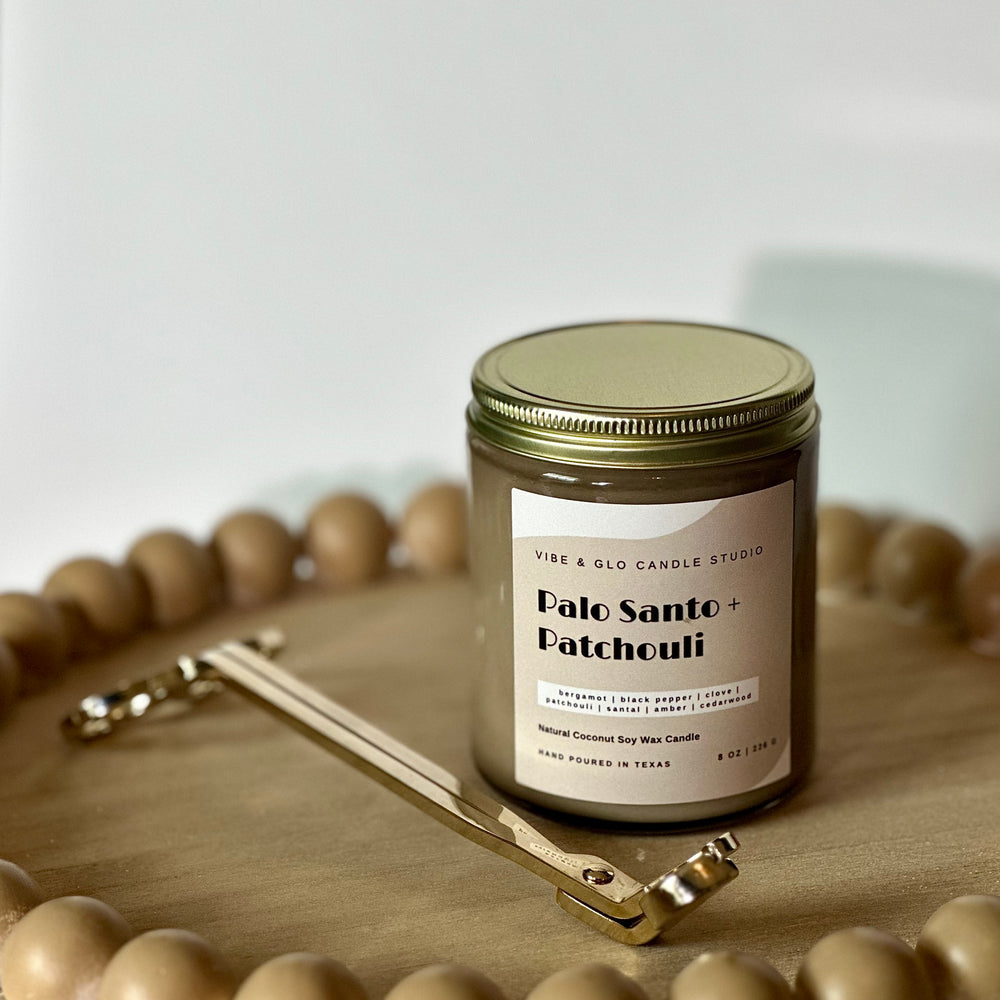 
                  
                    Vibe & Glo Candle Studio's Palo Santo + Patchouli candle styled with a gold wick trimmer on a beaded wooden tray. Features an elegant gray and white label showcasing its rich and grounding fragrance.
                  
                