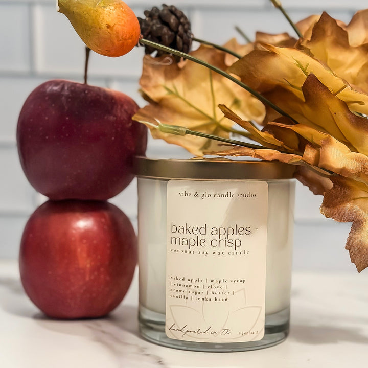 Candle labeled 'baked apples maple crisp' with apples and leaves on a marble surface