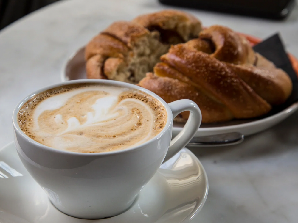 Close-up of a cappuccino with latte art served in a white cup, paired with a sweet pastry on a plate in the background — cozy coffee shop breakfast scene.