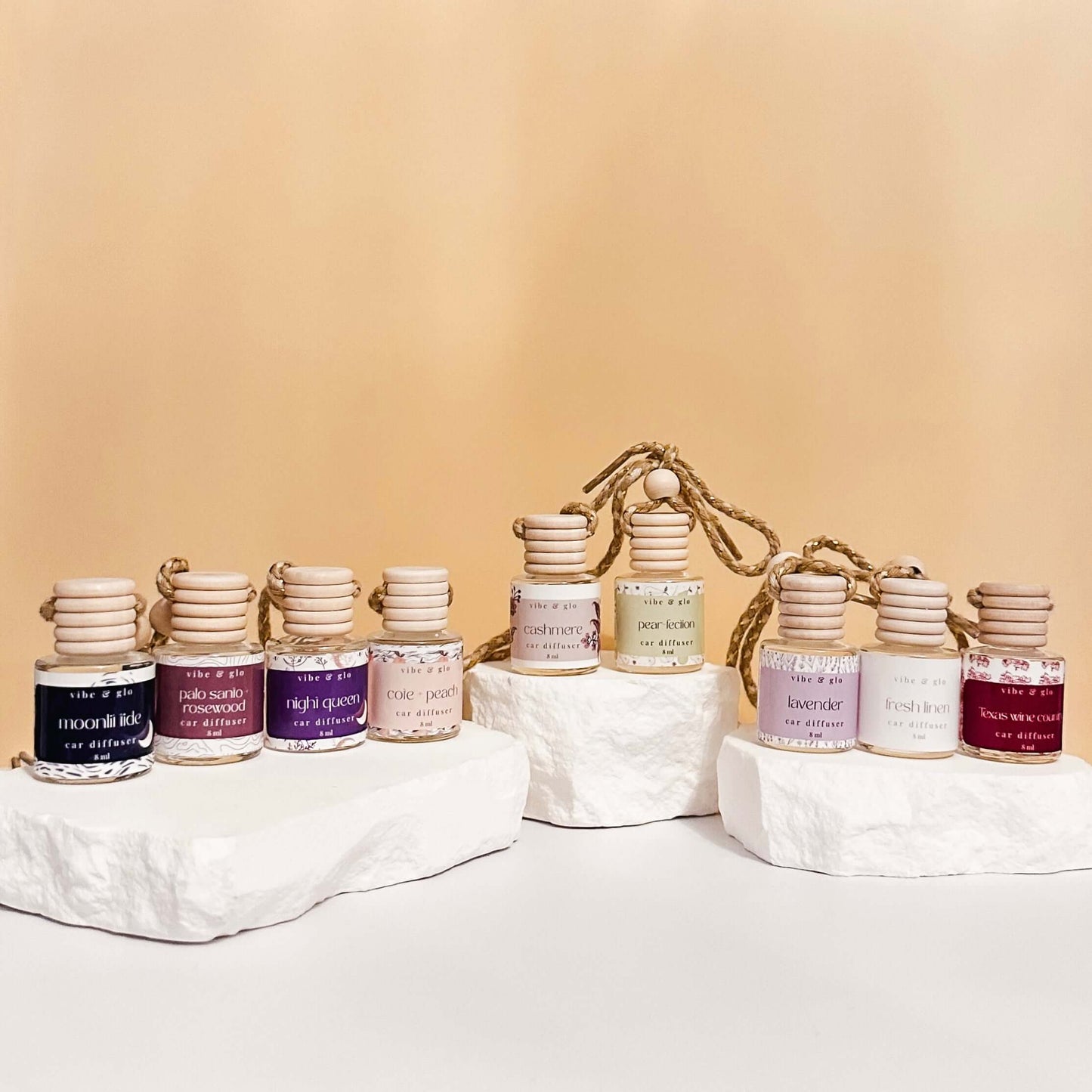 Assortment of refillable car air fresheners in elegant glass bottles displayed on white rocks, featuring various scents.