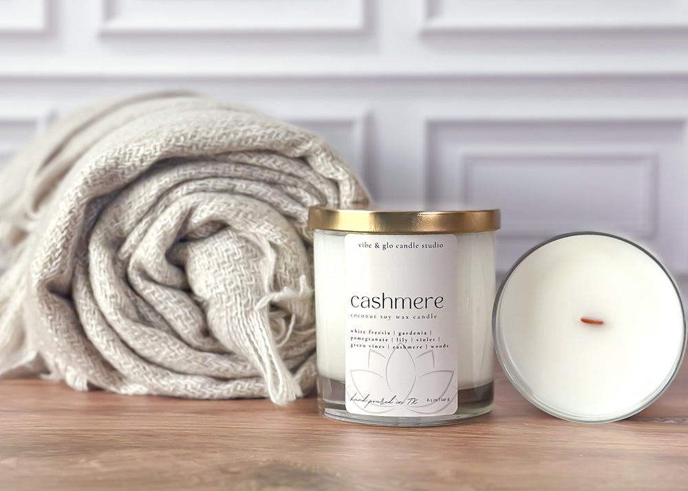 Candle labeled 'cashmere' next to a folded blanket on a wooden surface with a white paneled wall background.